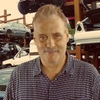 Doug Jordan Man with light brown hair and a salt-and-pepper mustache standing in front of an auto yard